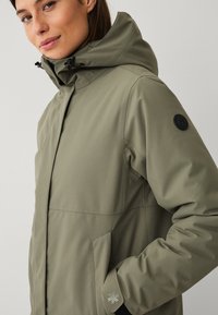 Donna che indossa una giacca invernale verde con cappuccio e colletto alto, con toggles neri regolabili e una patch nera rotonda sulla parte superiore della manica.