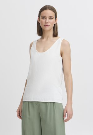 Egyenes barna hajú nő fehér ujjatlan felsőt és világoszöld, bő nadrágot visel, egy egyszerű világos háttér előtt állva.