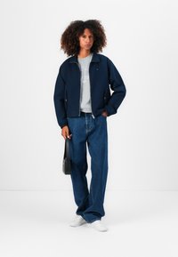 Marine bomberjack met ritssluiting en elastische manchetten, grijze sweatshirt eronder, losse blauwe jeans en witte sneakers. Zwarte handtas.