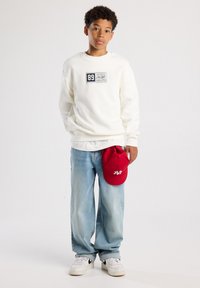 Sudadera blanca con un gráfico rectangular, jeans holgados de color azul claro y una gorra de béisbol roja sostenida en la mano. Zapatillas blancas completan el look.