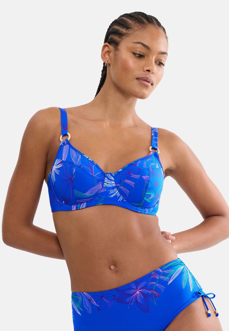 Donna che indossa un bikini blu con motivo floreale, spalline regolabili e dettagli con anelli dorati, posando con le mani dietro la schiena.
