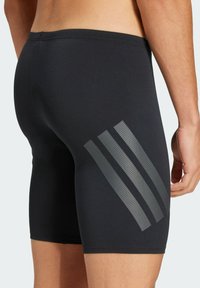 Pantalones cortos deportivos negros hechos de material elástico, con tres rayas grises con un patrón perforado en el lateral. Textura suave.