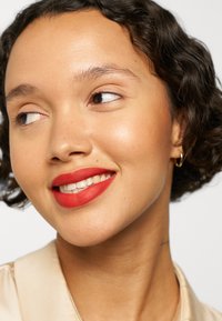 Close-up of a face featuring smooth skin, short curled hair, a warm beige top, and bold red lipstick, highlighting bright, even teeth.