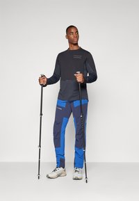 Long-sleeve black and gray athletic shirt, blue and navy pants with pockets and panels, sturdy trekking poles, light-colored shoes.