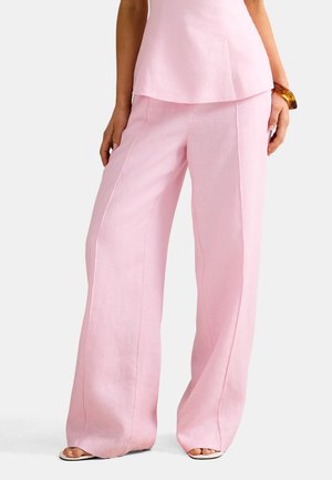 Naine, kes kannab heleroosasid laia säärega linaseid pükse sobiva varrukateta toppi ja kilpkonnakujulist käevõru, seisab käed lõdvestunult.