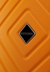 Valise orange texturée avec une plaque logo noire en forme de diamant portant l'inscription "WITTCHEN" en lettres dorées, présentant des stries angulaires pour le design.