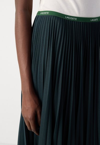 Jupe plissée vert foncé avec une ceinture de marque contrastante. Texture lisse et design fluide, avec des lignes verticales subtiles.