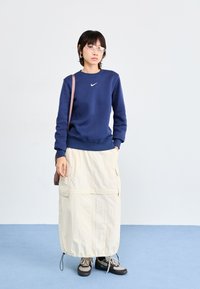 Σκούρο μπλε φούτερ με στρογγυλή λαιμόκοψη και λευκό λογότυπο Nike, σε συνδυασμό με μια κρέμα μακριά φούστα στιλ cargo και σκούρα αθλητικά παπούτσια.