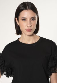 Camiseta negra con mangas acampanadas de ribete transparente y fruncido, cuello redondo y silueta ajustada. Tela suave con una textura delicada.