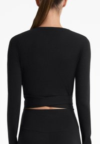 Black long-sleeve top, ribbed texture, cropped design with a knotted accent at the waist. Shown from the back against a white background.