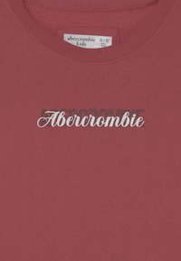 Rød bomuld T-shirt med teksten "Abercrombie" i hvid kursiv over mørke blokbogstaver. Rund hals med ribstrikket tekstur. Størrelse 11/12.