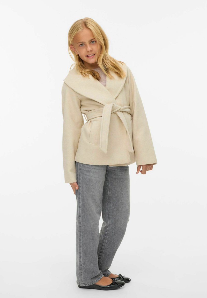 Bežinė apklijuota striukė su plačia šaliais apykakle, diržo liemeniu ir dviem priekinių kišenėmis, derinama su pilkomis tiesiomis kelnėmis ir juodomis butais.