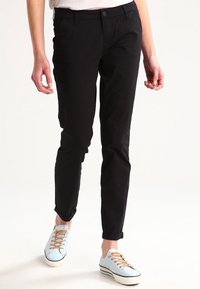 Black fitted trousers with a smooth texture, featuring side pockets and rolled-up cuffs, paired with light blue lace-up sneakers.