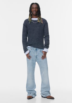 Dunkel gestreifter Strickpullover, hellblaue weite Jeans, braune Schuhe. Der Pullover hat einen lockeren Schnitt mit sichtbaren Rippungen und geschichteten Bündchen.