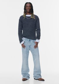 Temno prugasta pletena pulover, svetlo modri široki kavbojki, rjava obutev. Pulover ima ohlapen kroj z vidnim rebrastim vzorcem in slojevitimi manšetami.