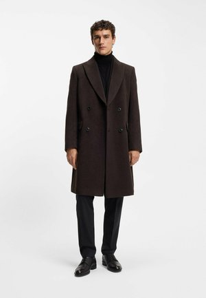 Manteau en laine brun avec des revers en pointe, design double boutonnage et boutons noirs. Porté sur un col roulé noir et un pantalon sombre.