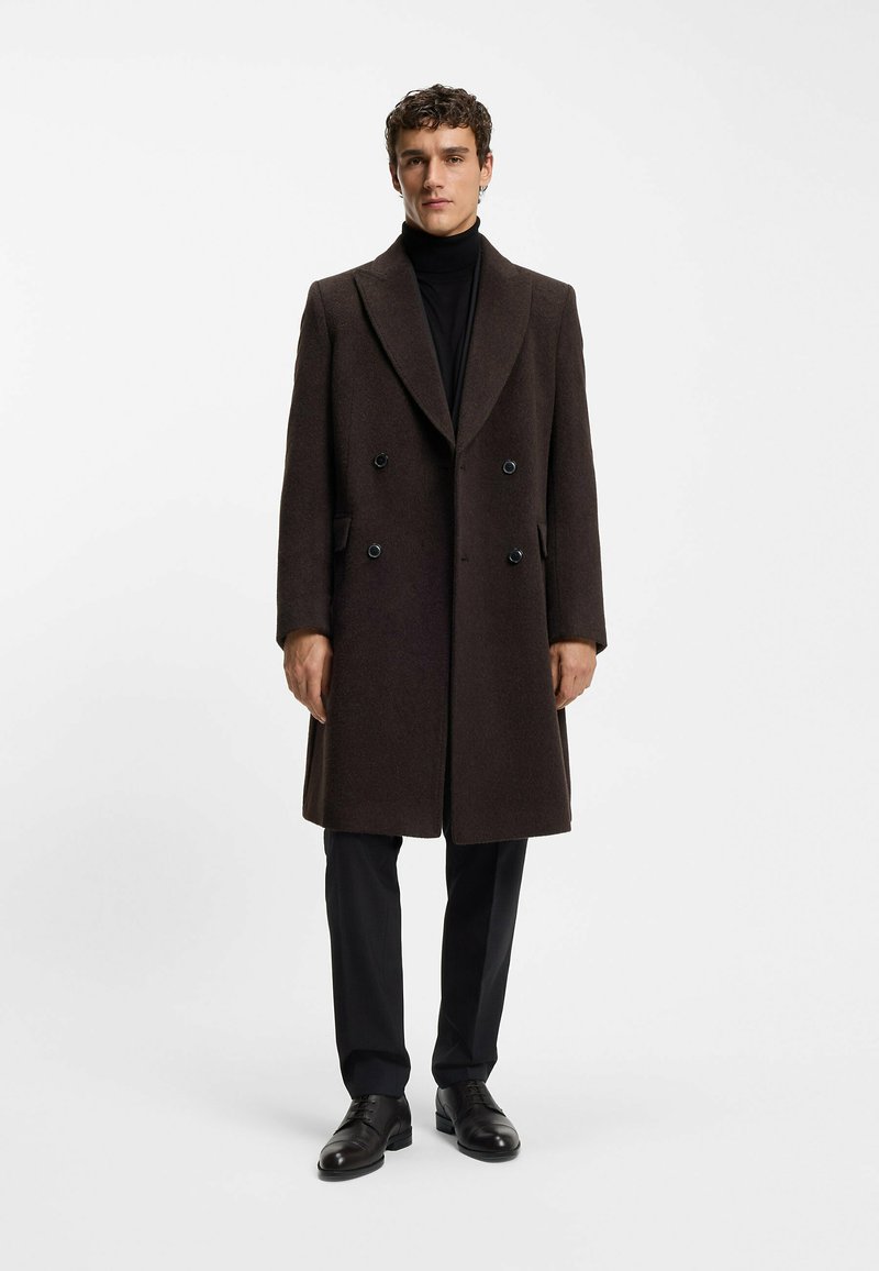 Cappotto di lana marrone con reverse a punta, design doppiopetto e bottoni neri. Indossato sopra un dolcevita nero e pantaloni scuri.