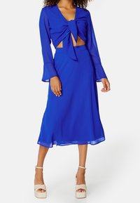 Femme portant un crop top à manches longues bleu royal avec un nœud devant et une jupe midi assortie, accompagnée de talons compensés beiges.