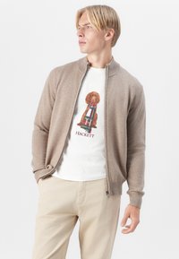 Bežna zip-up cardigan s rebrastim ovratnikom in manšeti, nosi se čez belo majico s grafikami rjavega psa. Svetlo bež hlače.