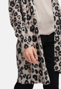 Cardigan so vzorom leopardích škvrn v mäkkej a chlpatej látke s odtieňmi béžovej a čiernej. Otvorený predok, dlhé rukávy, voľný strih a bez zapínania.