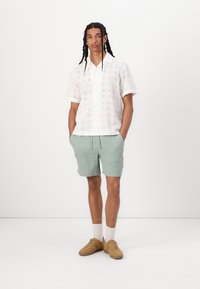 Weißes kurzärmliges Hemd mit Lochmuster, kombiniert mit mintgrünen Shorts. Braune Sneaker und weiße Socken runden den Look ab.