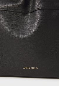 Fekete textúrájú bőrtáska, amely diszkrét hajtogatott dizájnnal és arany színű, dombornyomott "ANNA FIELD" márkanevével rendelkezik az alján.