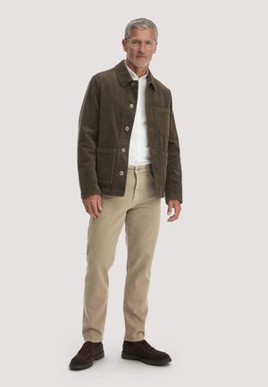 Mann mit Brille und grauen Haaren, brauner Knopfjacke, weißem Hemd, beiger Hose und dunkelbraunen Schuhen, stehend vor einfarbigem Hintergrund.