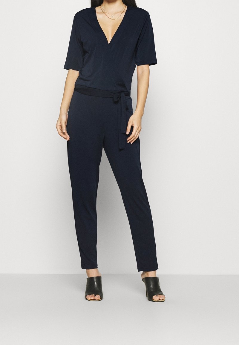 Combinaison bleu marine avec un décolleté en V plongeant, des manches courtes et une ceinture à nouer à la taille. Tissu lisse et design de jambe fuselée. Mules noires à bout ouvert.