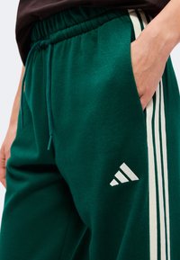 Εγγύς λήψη πράσινου παντελονιού Adidas με λευκές ρίγες στο πλάι και χέρι στην τσέπη.