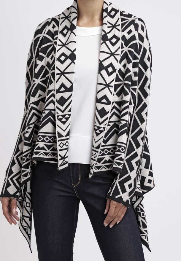 Chal negro y beige con patrones geométricos, abertura frontal, mangas largas y textura de punto suave, combinado con una camisa blanca y vaqueros oscuros.