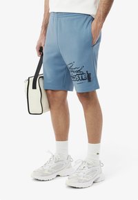 Bermudas de algodón azul con un estampado del logo negro de Lacoste. Combinadas con zapatillas deportivas blancas y un bolso beige con detalles negros.