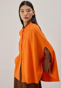 Cape en maille orange avec un col montant, des boutons sur le devant et des fentes aux manches. Portée par-dessus un vêtement marron. Texture lisse, design structuré.