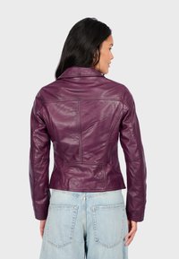 Fialová kožená bunda s přiléhavým střihem, viditelnými detaily šití a klasickým límcem. Nosená přes džínsy se širokými nohavicemi a světle modrou barvou.