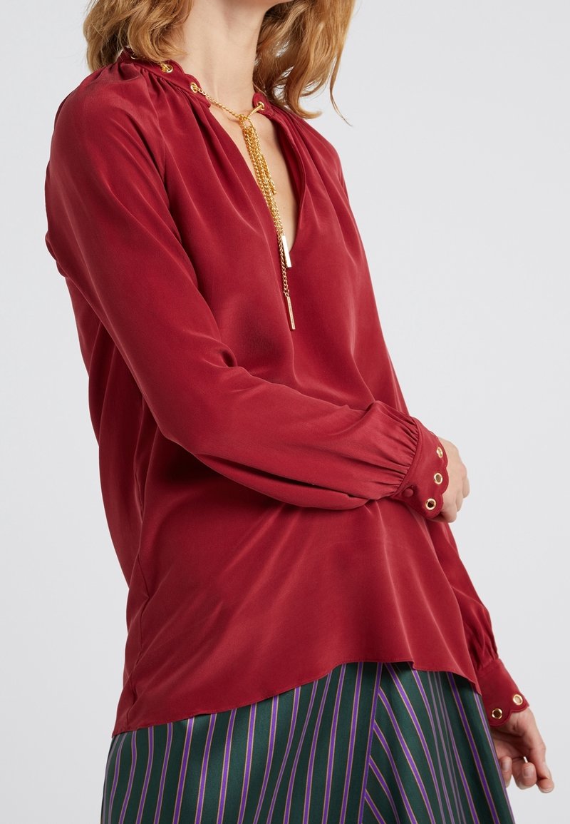 Femme portant un chemisier en satin rouge à manches longues avec des boutons dorés et un collier chaîne doré, associé à une jupe rayée.