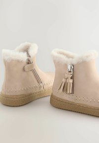 Beige ankelstøvler lavet af blødt materiale med for af faux pels, sidelukning og dekorative frynser, placeret på en struktureret, lysfarvet sål.