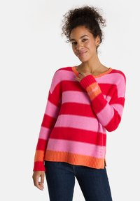 Frau mit lockigem Haar, die einen gestreiften Pullover in Rot, Pink und Orange trägt, lächelt und berührt mit ihrer rechten Hand den Halsausschnitt des Pullovers.