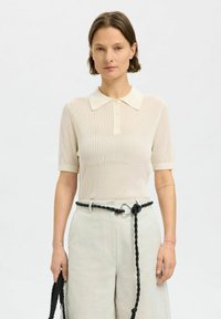 Polo en tricot côtelé crème avec des manches courtes et un col, doté d'une fermeture à boutons et d'une texture douce. Associé à des pantalons clairs taille haute.