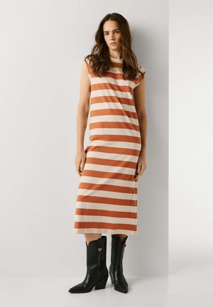 Femme debout contre un mur blanc, portant une robe sans manches longueur genou à rayures horizontales beige et rouille, ainsi que des bottes noires mi-mollet.