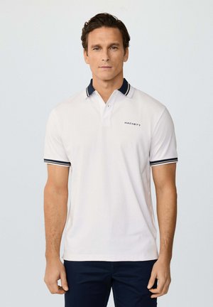 Hombre con camiseta tipo polo blanca de Hackett, con ribetes azul marino en el cuello y las mangas, posando frente a un fondo claro y liso.