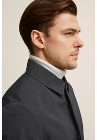 Musta kauluspuku, napitettu, sujuvalla pinnalla. Kerroksittain vaaleanharmaan poolopaidan päällä. Profiilinäkymä neutraalia taustaa vastaan.