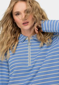 Niebieski i kremowy sweter w paski z suwakiem przy kołnierzu, o ribbanym wykończeniu, luźnym kroju i okrągłym srebrnym uchwycie suwaka.