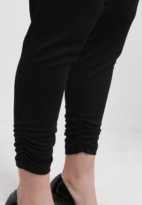 Czarne legginsy z marszczonym detalem przy kostce, wykonane z elastycznego materiału, połączone z błyszczącymi czarnymi szpilkami.