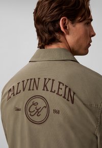 Νέος άνδρας που φοράει χακί μπουφάν με "CALVIN KLEIN Since 1968" και κυκλικό λογότυπο CK κεντημένο στην πλάτη, στραμμένος πλάγια.