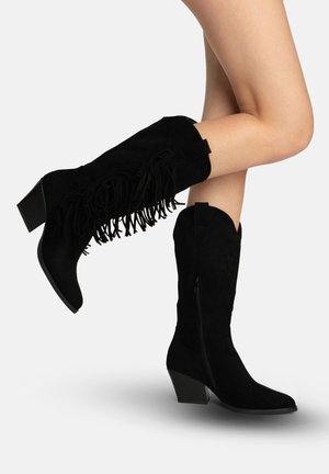 Botas negras de ante hasta la mitad de la pantorrilla con tacones apilados y detalles de flecos, usadas en piernas desnudas cruzadas sobre un fondo liso.