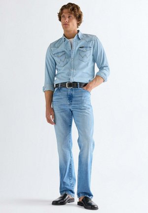 Mężczyzna w jasnoniebieskiej koszuli dżinsowej i dżinsach, czarnym pasku oraz czarnych butach wsuwanych, stojący z jedną ręką w kieszeni na białym tle.