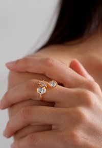 Goldener Ring, besetzt mit drei klaren, tropfenförmigen Steinen, zwei größeren auf einer Seite. Hand, die das Handgelenk greift, mit weicher Hautstruktur im Hintergrund.