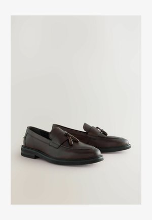 Mocasines de cuero marrón con suela plana negra, que cuentan con una tira decorativa en el frontal y borlas. Textura suave con detalles de costura sutiles.