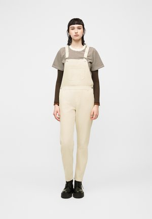 Ung person som står rett opp og ned, iført beige overalls, stripet kortermet skjorte over brun langermet trøye, og svarte snørestøvler.