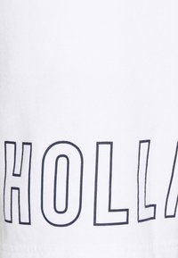 Vit t-shirt med fet marinblå text som stavar "HOLLAND." Tyget verkar mjukt med en slät yta och klassisk rund halsringning.