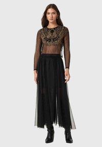 Haut noir transparent à manches longues avec un design orné, assorti à une jupe en tulle noir fluide. Modèle portant des bottes noires jusqu'aux genoux.